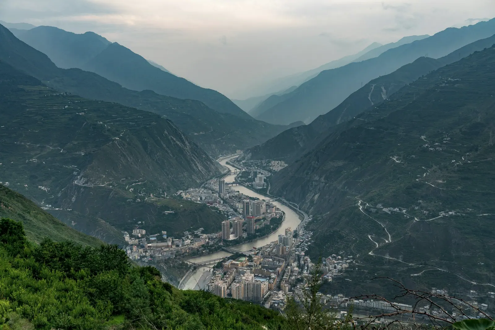 A river cutting through a deep green gorge, a small town on the slope wrapped in mist