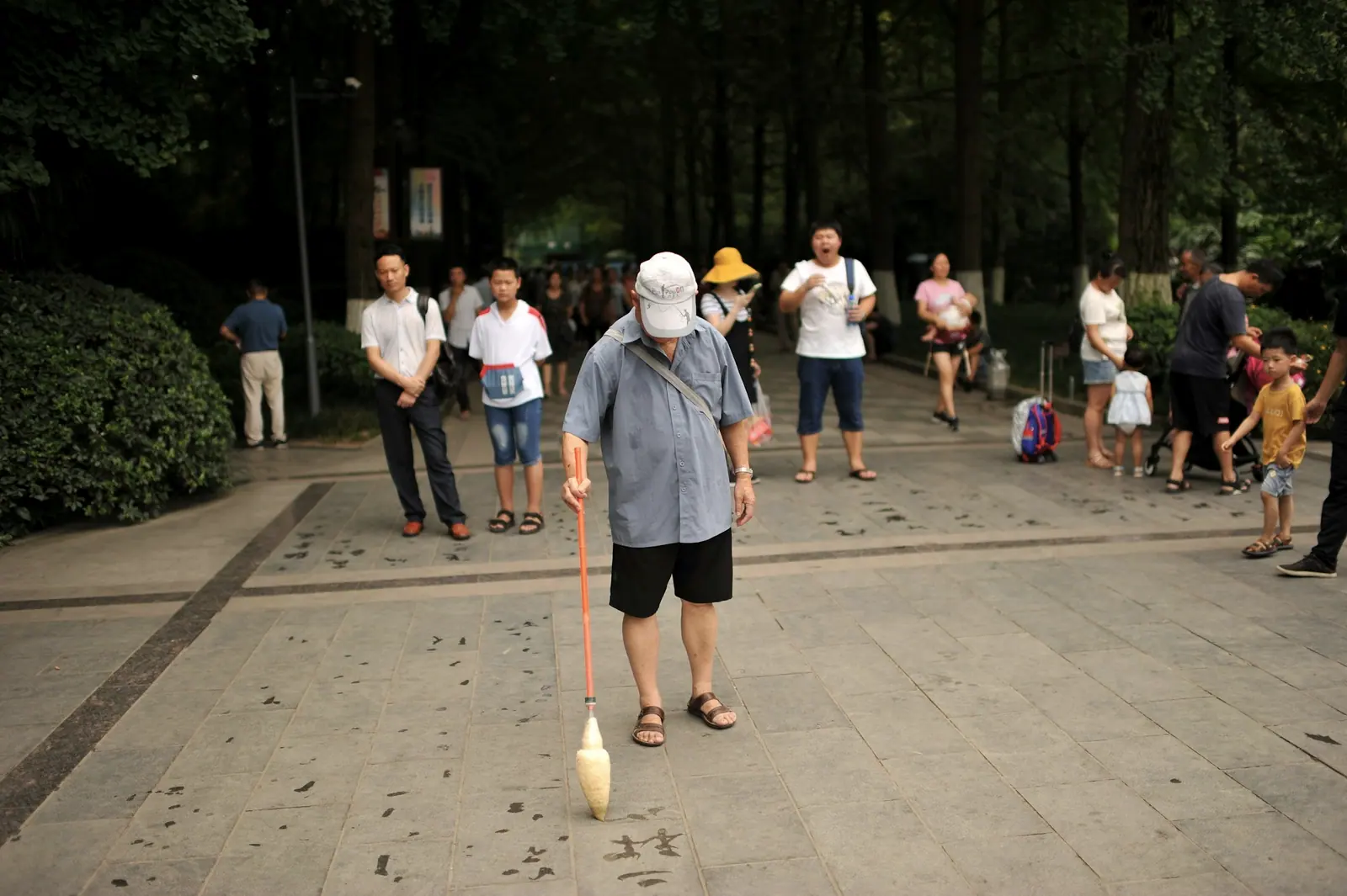 An old man writing with water on the paving stones using a long brush, a few onlookers gathered around
