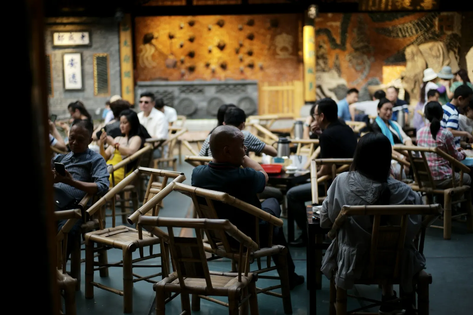 Customers sitting in bamboo chairs in an old teahouse, drinking tea and talking, most of them facing away from the camera
