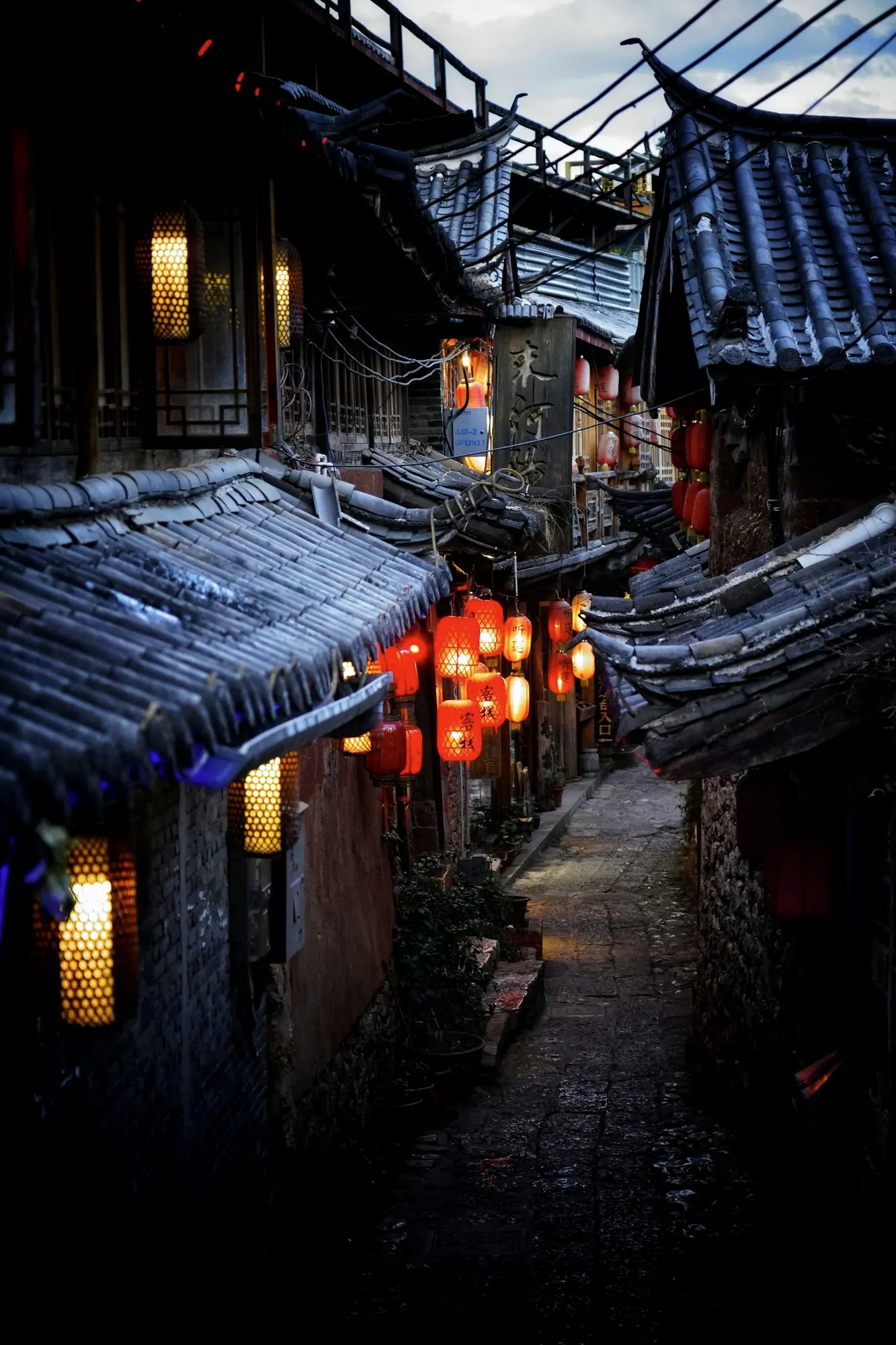 A narrow flagstone lane between grey-tiled eaves, the red lanterns already lit