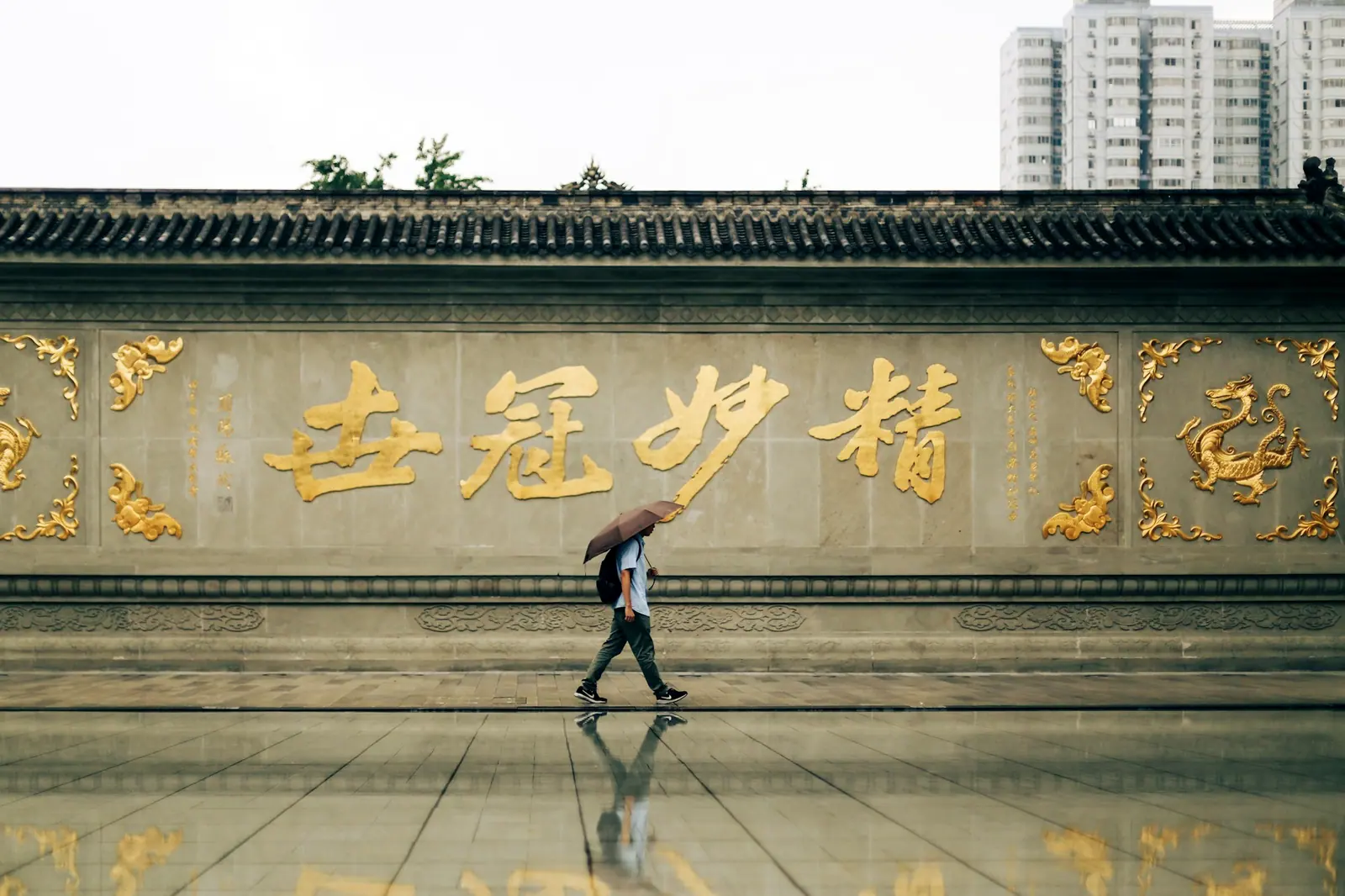 A person with an umbrella walks past a gilded carved screen wall, reflected in the puddle underfoot